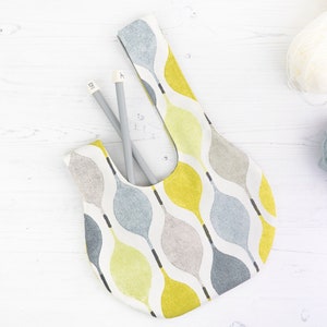 Puede incluir: Una bolsa de tela blanca con un patr&oacute;n abstracto amarillo, gris y azul. La bolsa est&aacute; sobre una superficie de madera blanca con dos agujas de tejer dentro.
