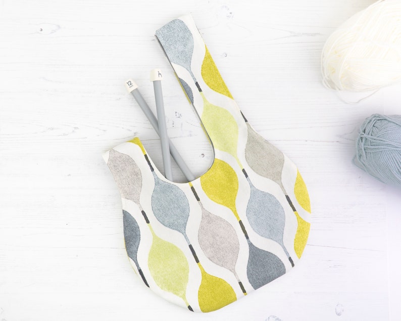 Puede incluir: Una bolsa de tela blanca con un patr&oacute;n abstracto amarillo, gris y azul. La bolsa est&aacute; sobre una superficie de madera blanca con dos agujas de tejer dentro.