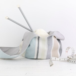 Puede incluir: Una bolsa de tela a rayas con forro gris, llena de una bola de hilo blanco y dos agujas de tejer grises. Una cinta m&eacute;trica blanca est&aacute; sobre una superficie de madera blanca.