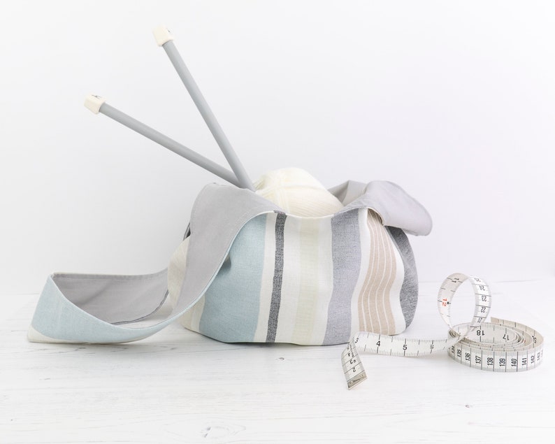 Puede incluir: Una bolsa de tela a rayas con forro gris, llena de una bola de hilo blanco y dos agujas de tejer grises. Una cinta m&eacute;trica blanca est&aacute; sobre una superficie de madera blanca.