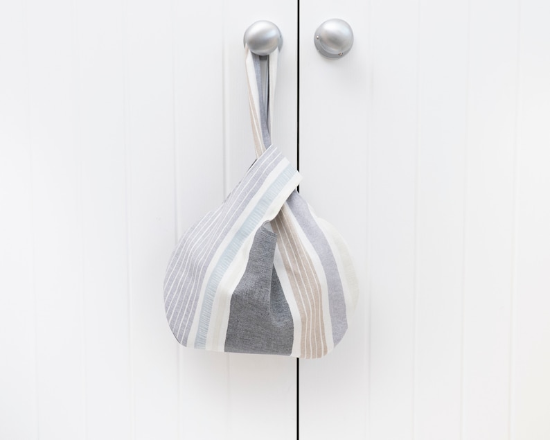 Puede incluir: Una bolsa de tela a rayas grises y blancas con un asa anudada. La bolsa est&aacute; colgada de una puerta blanca.