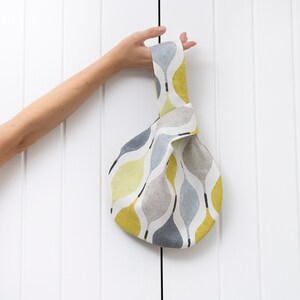 Puede incluir: Una bolsa de tela anudada con un patr&oacute;n abstracto gris, blanco y amarillo. La bolsa tiene un solo asa y est&aacute; hecha de una tela suave y tejida.