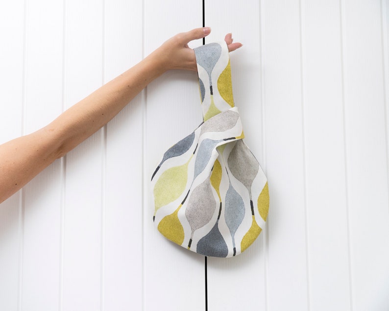 Puede incluir: Una bolsa de tela anudada con un patr&oacute;n abstracto gris, blanco y amarillo. La bolsa tiene un solo asa y est&aacute; hecha de una tela suave y tejida.