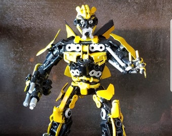 May include: A yellow and black robot made of metal nuts, bolts, and other hardware. The robot has a human-like face and is posed in a standing position.
