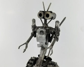 May include: A metal robot sculpture, handcrafted from various metal components. The robot has a head with two cylindrical eyes, antennae, and articulated arms with claw-like hands. The body is made from metal parts, including a chain-link base. The text "S.E.C. I.M.C.3/4" is visible.