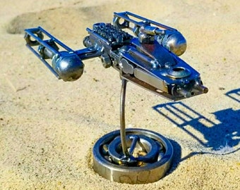May include: A detailed metal spaceship sculpture, crafted from various metallic parts, standing on a bearing base. The artwork is displayed on a sandy beach, set against a clear blue sky, showcasing intricate design.
