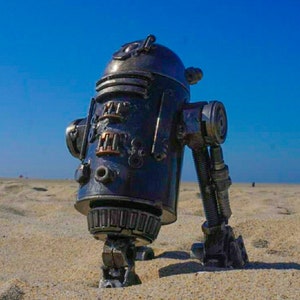 May include: A robot made of metal scraps, standing on a sandy beach with a blue sky in the background.