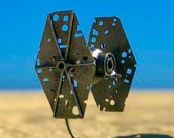 May include: A metal sculpture of a TIE fighter from Star Wars. The sculpture is made of cut and welded metal and is painted in a silver colour. The sculpture is sitting on a sandy surface.