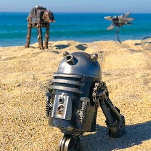 Puede incluir: Una figurita de robot de metal al estilo de R2-D2 de Star Wars, de pie en una playa de arena. El robot está hecho de piezas de metal recicladas y tiene un aspecto desgastado e industrial. En el fondo, se pueden ver otras dos figuritas de metal, un caminante y una nave espacial, en la playa.