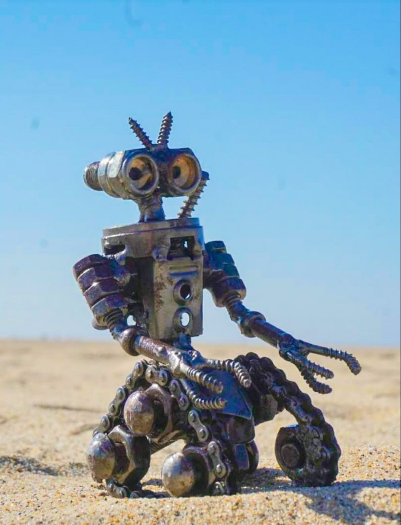 Puede incluir: Un robot hecho de piezas met&aacute;licas, incluyendo tuercas, tornillos y cadenas. El robot tiene una forma humanoide con dos brazos, dos piernas y una cabeza con dos ojos grandes. El robot est&aacute; sentado en una superficie arenosa.