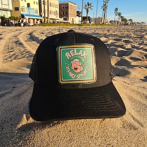 Op de afbeelding: Zwarte truckerpet met een vierkante patch met Popeye de zeeman. De patch heeft een groene achtergrond met de woorden "RELAX, IT'S ONLY SPINACH" en een afbeelding van Popeye. De pet ligt op een zandstrand.