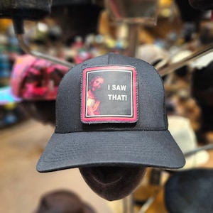 May include: Black trucker hat with a curved brim and mesh back. The hat features a rectangular patch with an image of Jesus and the text "I SAW THAT!" in white letters on a black background with a red border.
