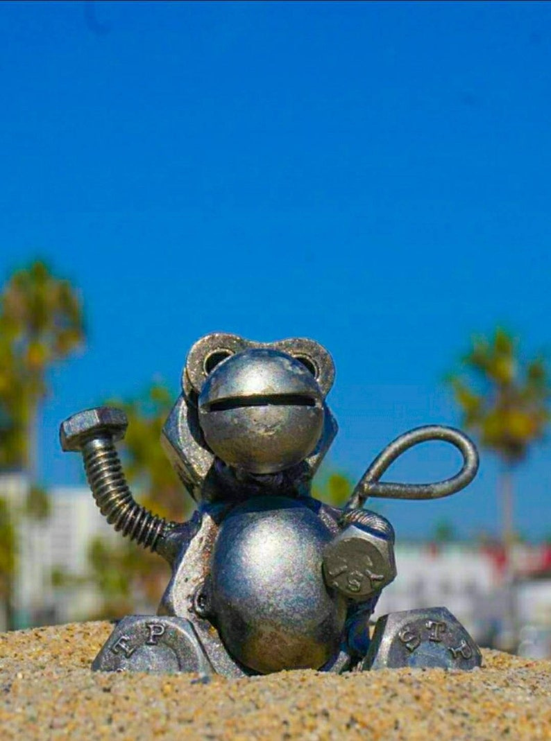 May include: A silver metal sculpture of a frog sitting on a sandy surface. The frog is made of nuts, bolts, and other metal pieces. The frog has a large head with a wide mouth and a small body. The frog's arms are raised in the air, and its legs are bent at the knees.