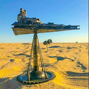 Puede incluir: Una escultura metálica de una nave espacial, con un diseño detallado, se alza sobre una base de trípode. La escultura se encuentra sobre arena, bajo un cielo azul claro, con otro objeto metálico más pequeño.