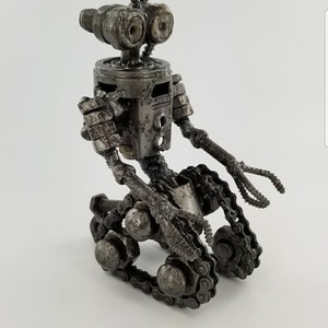 Puede incluir: Una peque&ntilde;a escultura de robot de metal hecha de tuercas, tornillos y cadenas. El robot tiene una cabeza redonda con dos ojos grandes, un cuerpo peque&ntilde;o y dos brazos con manos. El robot est&aacute; sentado sobre un par de orugas.