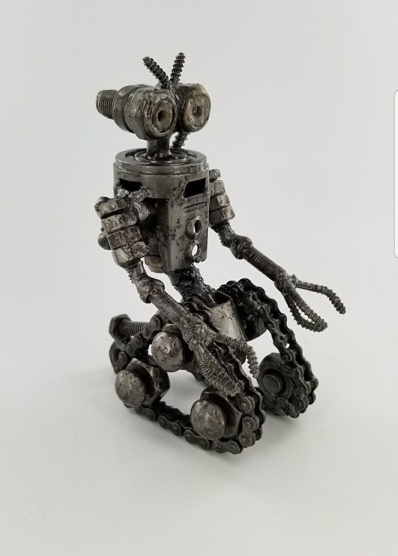 Puede incluir: Una peque&ntilde;a escultura de robot de metal hecha de tuercas, tornillos y cadenas. El robot tiene una cabeza redonda con dos ojos grandes, un cuerpo peque&ntilde;o y dos brazos con manos. El robot est&aacute; sentado sobre un par de orugas.