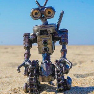 Puede incluir: Un robot hecho de piezas met&aacute;licas, incluyendo tuercas, tornillos y cadenas. El robot tiene un cuerpo azul, una cabeza plateada y dos ojos grandes y redondos. El robot est&aacute; sentado en una superficie arenosa.