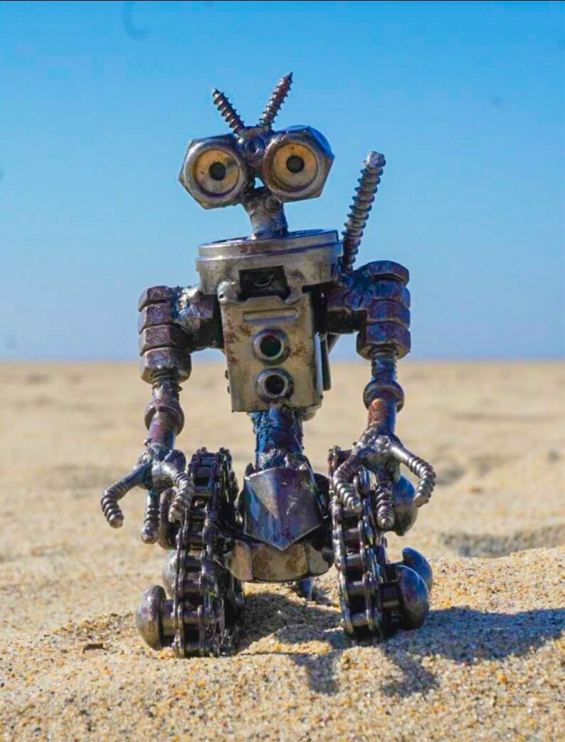 Puede incluir: Un robot hecho de piezas met&aacute;licas, incluyendo tuercas, tornillos y cadenas. El robot tiene un cuerpo azul, una cabeza plateada y dos ojos grandes y redondos. El robot est&aacute; sentado en una superficie arenosa.