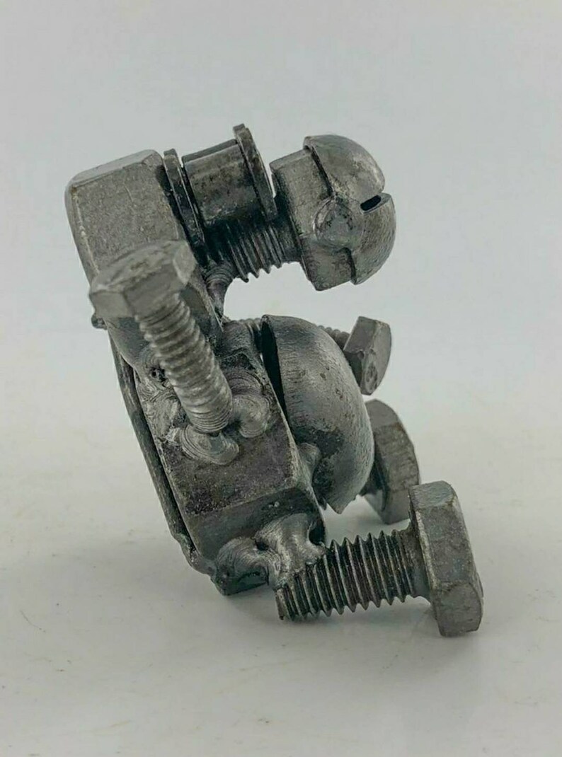 May include: A small, abstract sculpture made from metal nuts and bolts. The sculpture is in the shape of a person sitting down, with the head tilted to the side. The sculpture is made of a silver metal.