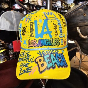 Könnte beinhalten: Gelbe Baseballkappe mit den Aufschriften "LA LOS ANGELES" und "VENICE BEACH" in Blau, Grün und Orange. Die Kappe hat eine blaue Kordel und weitere farbige Texte und Grafiken.