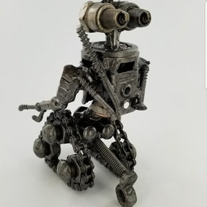 Puede incluir: Una figura de robot hecha de piezas de metal recicladas, incluyendo tuercas, tornillos y cadenas. El robot tiene un cuerpo plateado y ojos negros hechos de buj&iacute;as. El robot est&aacute; de pie sobre una superficie blanca.