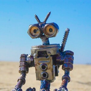 Puede incluir: Un robot hecho de metal y materiales reciclados, con grandes ojos redondos y un acabado oxidado. El robot est&aacute; sentado con las piernas cruzadas y los brazos apoyados en las rodillas.