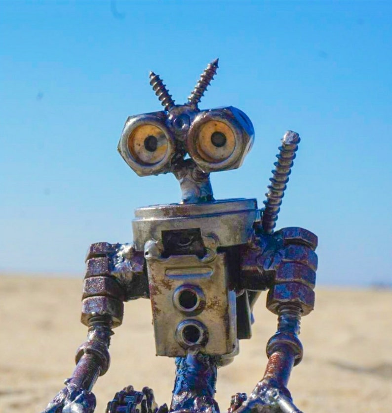 Puede incluir: Un robot hecho de metal y materiales reciclados, con grandes ojos redondos y un acabado oxidado. El robot est&aacute; sentado con las piernas cruzadas y los brazos apoyados en las rodillas.