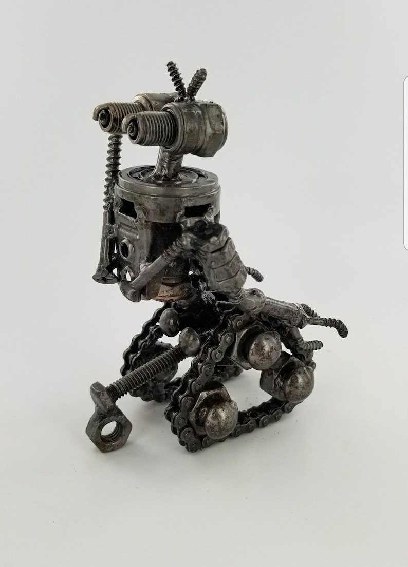 Puede incluir: Una peque&ntilde;a escultura de robot hecha de piezas de metal recicladas. La escultura est&aacute; hecha de tuercas, tornillos, tornillos y otras piezas de metal. El robot tiene un sistema de orugas de cadena y una cabeza hecha de buj&iacute;as.