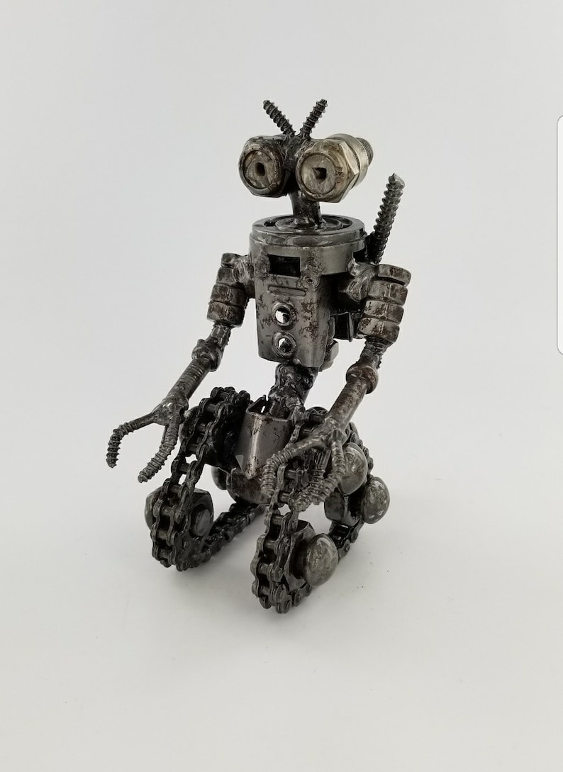 Puede incluir: Una peque&ntilde;a escultura de robot de metal hecha con materiales reciclados. El robot est&aacute; sentado con las piernas cruzadas y tiene un sistema de orugas de cadena para sus pies. El robot tiene una cabeza grande con dos ojos grandes y una peque&ntilde;a antena.