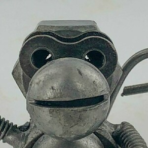 May include: A metal sculpture of a monkey made from recycled metal parts. The monkey has a large head with a round face and large eyes. The monkey is holding a wrench in one hand and a hook in the other.