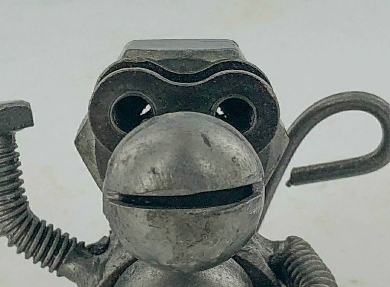 May include: A metal sculpture of a monkey made from recycled metal parts. The monkey has a large head with a round face and large eyes. The monkey is holding a wrench in one hand and a hook in the other.