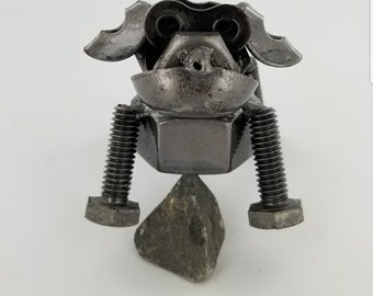May include: A metal sculpture of a dog-like creature made from nuts, bolts, and other hardware. The sculpture has a grey colour and is balanced on a small rock. The dog has a whimsical, industrial design.