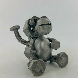 May include: A small, grey metal sculpture of a robot made from nuts and bolts. The robot is sitting with one arm raised and has a round head with two large eyes.