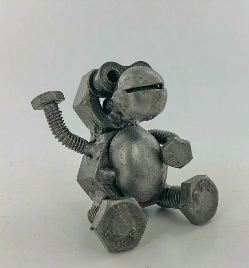 May include: A small, grey metal sculpture of a robot made from nuts and bolts. The robot is sitting with one arm raised and has a round head with two large eyes.