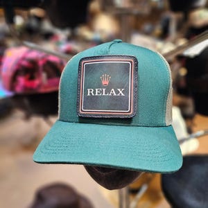 Op de afbeelding: Een groene truckerpet met een mesh achterkant en een gebogen klep. De voorkant heeft een rechthoekige patch met het woord "RELAX" en een kroongrafiek. De pet wordt gepresenteerd tegen een wazige achtergrond van andere petten.