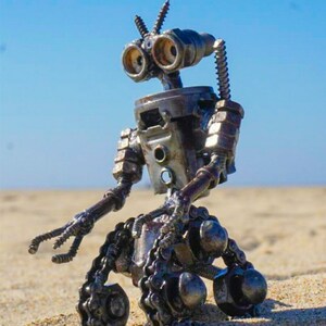 Puede incluir: Un robot hecho de piezas met&aacute;licas, incluyendo tuercas, tornillos y cadenas, est&aacute; sentado en una superficie arenosa. El robot tiene una cabeza redonda con dos ojos grandes y dos antenas. El robot tiene dos brazos y dos piernas, y su cuerpo est&aacute; hecho de varias piezas met&aacute;licas.