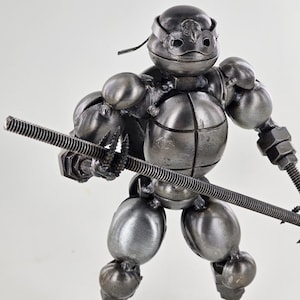 May include: A metal sculpture of a Teenage Mutant Ninja Turtle holding a weapon. The sculpture is made of nuts, bolts, and other metal pieces. The turtle is wearing a mask and has a shell.