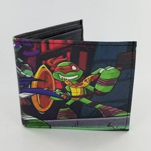 Könnte beinhalten: Ein zweifach gefaltetes Portemonnaie mit einer farbenfrohen Grafik der Teenage Mutant Ninja Turtles. Das Portemonnaie hat ein schwarzes Innenfutter und ein lebendiges Außendesign mit den Charakteren in Actionposen. Das Portemonnaie ist aus strapazierfähigem Material gefertigt.