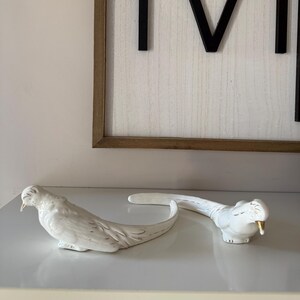 May include: Two white ceramic bird figurines with long, curved tails and gold-colored beaks. The birds are positioned on a white surface, with a framed wooden sign in the background. The sign has a white background and black letters.