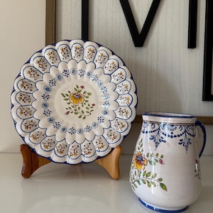 May include: A decorative ceramic plate and pitcher set. The plate is white with a scalloped edge, blue trim, and a floral design. The pitcher is white with blue floral patterns and a blue handle.