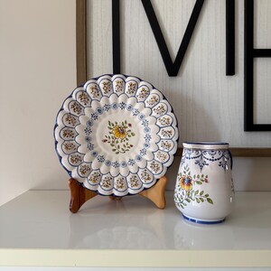 May include: A decorative ceramic plate with a scalloped edge, featuring a floral design in yellow, blue, and brown. A matching ceramic pitcher with a handle and similar floral motifs is also present. Both items are on a white surface.