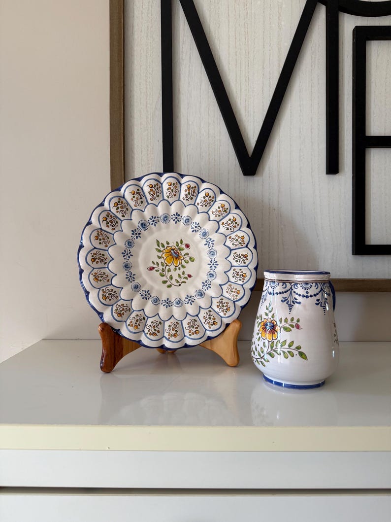 May include: A decorative ceramic plate with a scalloped edge, featuring a floral design in yellow, blue, and brown. A matching ceramic pitcher with a handle and similar floral motifs is also present. Both items are on a white surface.