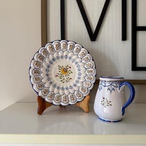 May include: A white ceramic plate and mug set with blue and yellow floral designs. The plate has a scalloped edge and sits on a wooden stand. The mug has a blue handle and rim, and both items have a vintage aesthetic.