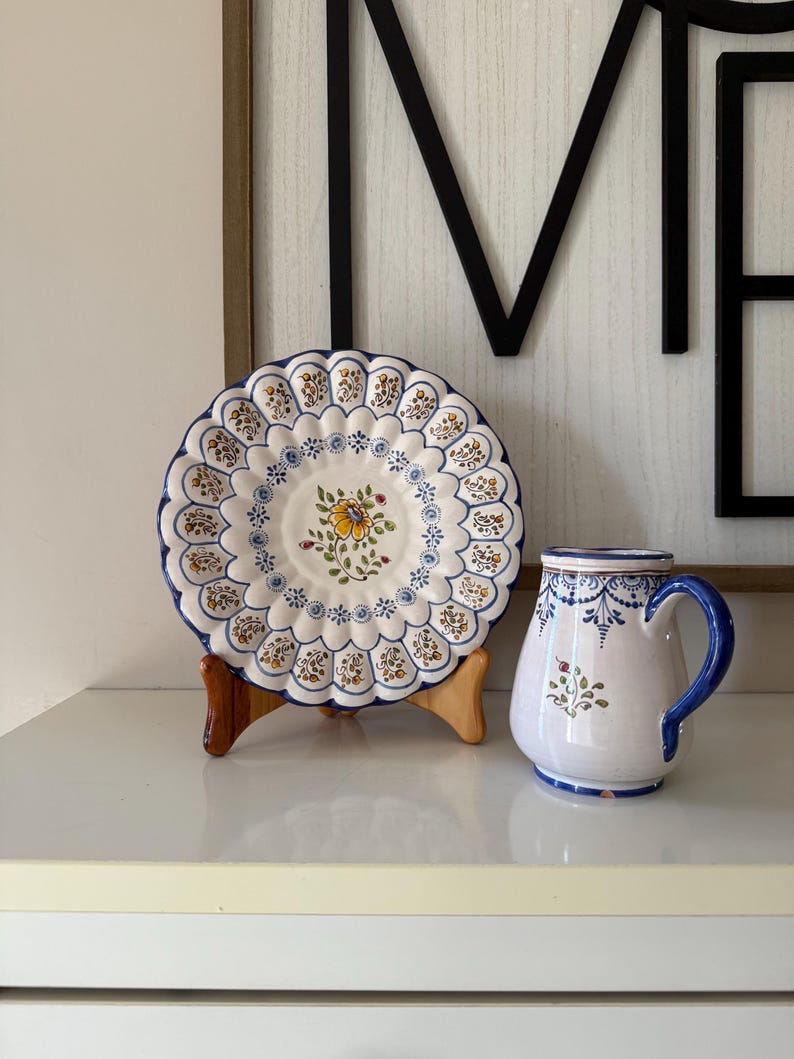 May include: A white ceramic plate and mug set with blue and yellow floral designs. The plate has a scalloped edge and sits on a wooden stand. The mug has a blue handle and rim, and both items have a vintage aesthetic.