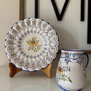 May include: A white ceramic plate with a scalloped edge and a floral design, displayed on a wooden stand. A matching pitcher with a handle and floral pattern sits beside the plate. Both items have blue and yellow accents.