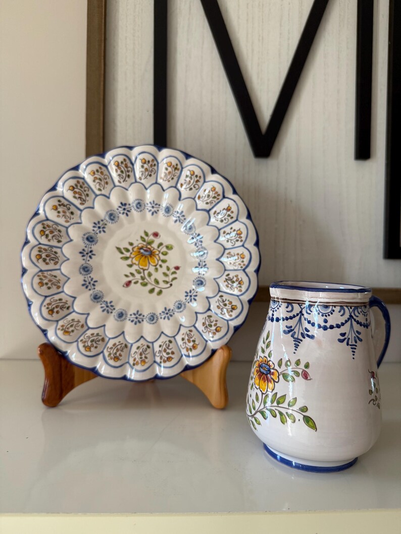 May include: A white ceramic plate with a scalloped edge and a floral design, displayed on a wooden stand. A matching pitcher with a handle and floral pattern sits beside the plate. Both items have blue and yellow accents.