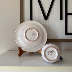 May include: A white ceramic plate and a small cup with blue accents. The plate is on a wooden stand. The bottom of the plate and cup have text and the number 1993. The background includes a framed black metal sign.