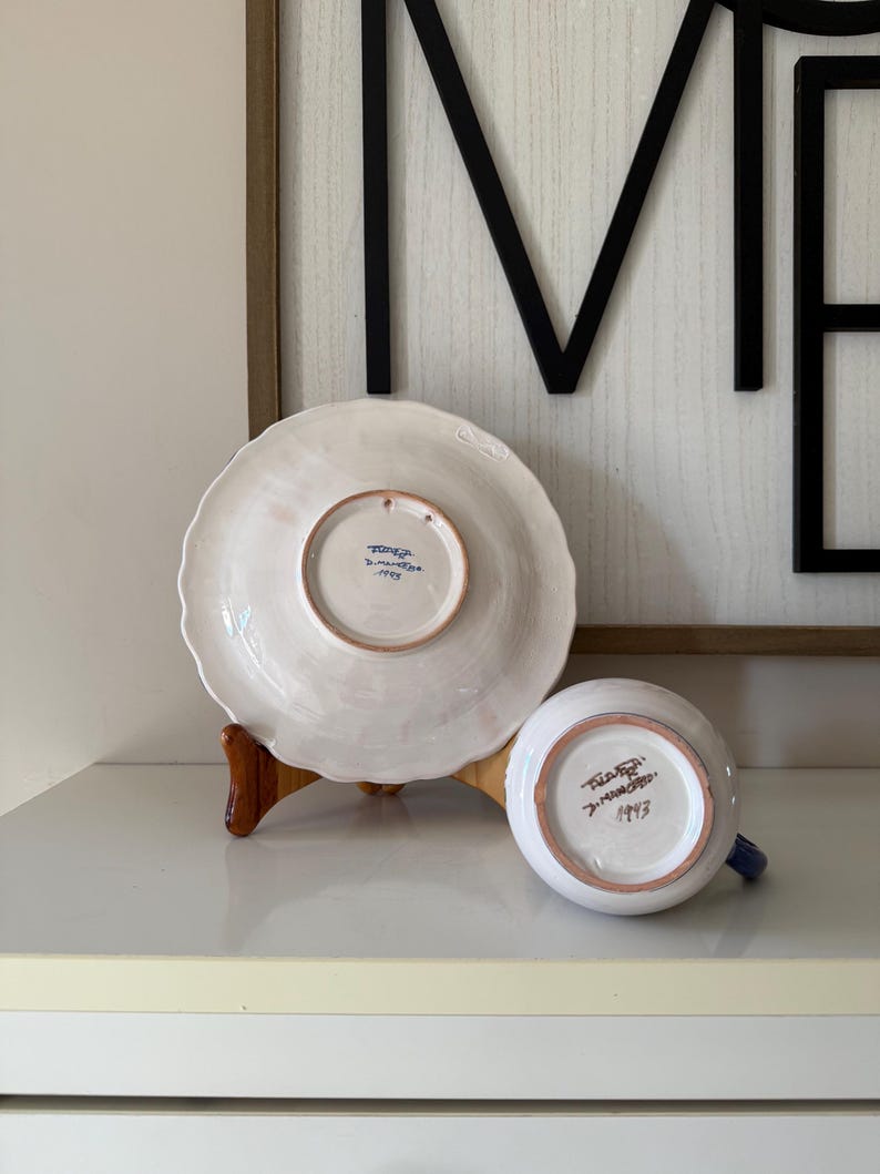 May include: A white ceramic plate and a small cup with blue accents. The plate is on a wooden stand. The bottom of the plate and cup have text and the number 1993. The background includes a framed black metal sign.