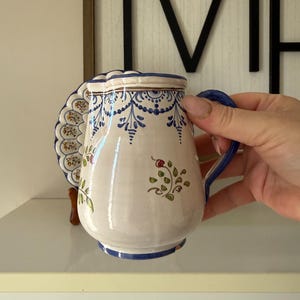 May include: A white ceramic pitcher with blue floral patterns and a matching handle. The pitcher has a scalloped edge and a decorative plate attached to the side. The design includes blue and brown accents, with a floral motif.