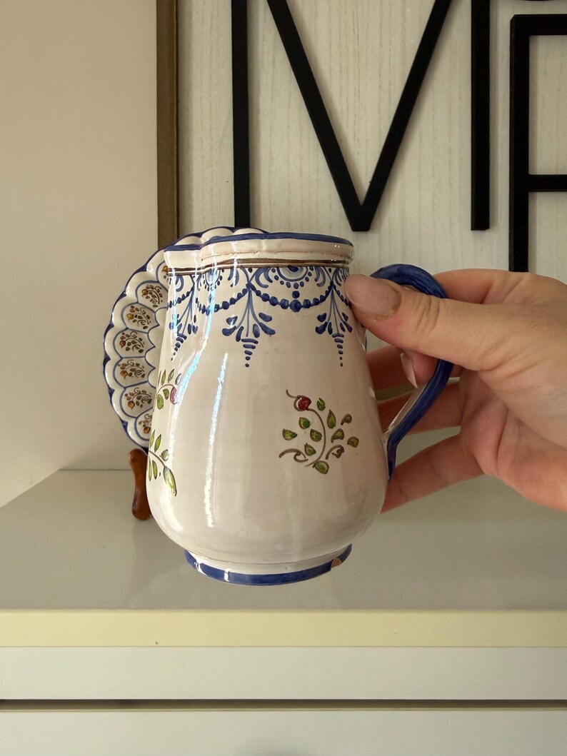 May include: A white ceramic pitcher with blue floral patterns and a matching handle. The pitcher has a scalloped edge and a decorative plate attached to the side. The design includes blue and brown accents, with a floral motif.